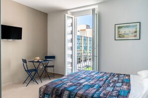 Ein Schlafzimmer mit einem Bett, einem an der Wand befestigten Fernseher, einem Balkon mit Blick auf Gebäude und einem kleinen Tisch mit Stühlen.