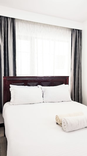 Una cama bien hecha con sábanas y almohadas blancas, un cabecero de madera y una toalla blanca enrollada al lado.