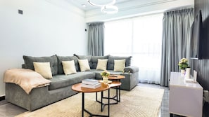 Una sala de estar moderna con un sofá gris, dos mesas redondas, un armario blanco y un amplio ventanal con cortinas.