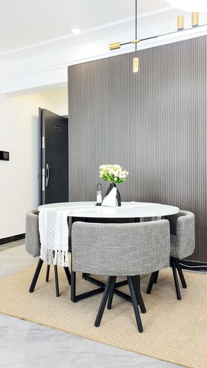Ein moderner Essbereich mit einem runden Tisch, grau gepolsterten Stühlen, einer Vase mit weißen Blumen und einer strukturierten Wand im Hintergrund.