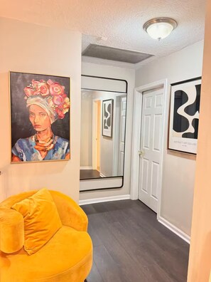 A hallway with a yellow chair, a painting, and a door leading to a room.