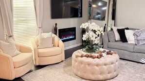 A modern living room with a fireplace, two armchairs, a sofa, a round ottoman, and a flat-screen TV. - Wellington Polo Tranquility (Wellington)