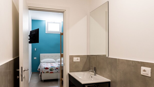 Un pasillo estrecho conduce a un dormitorio con cama y mesita de noche. A la derecha se ve un baño con lavabo y espejo.