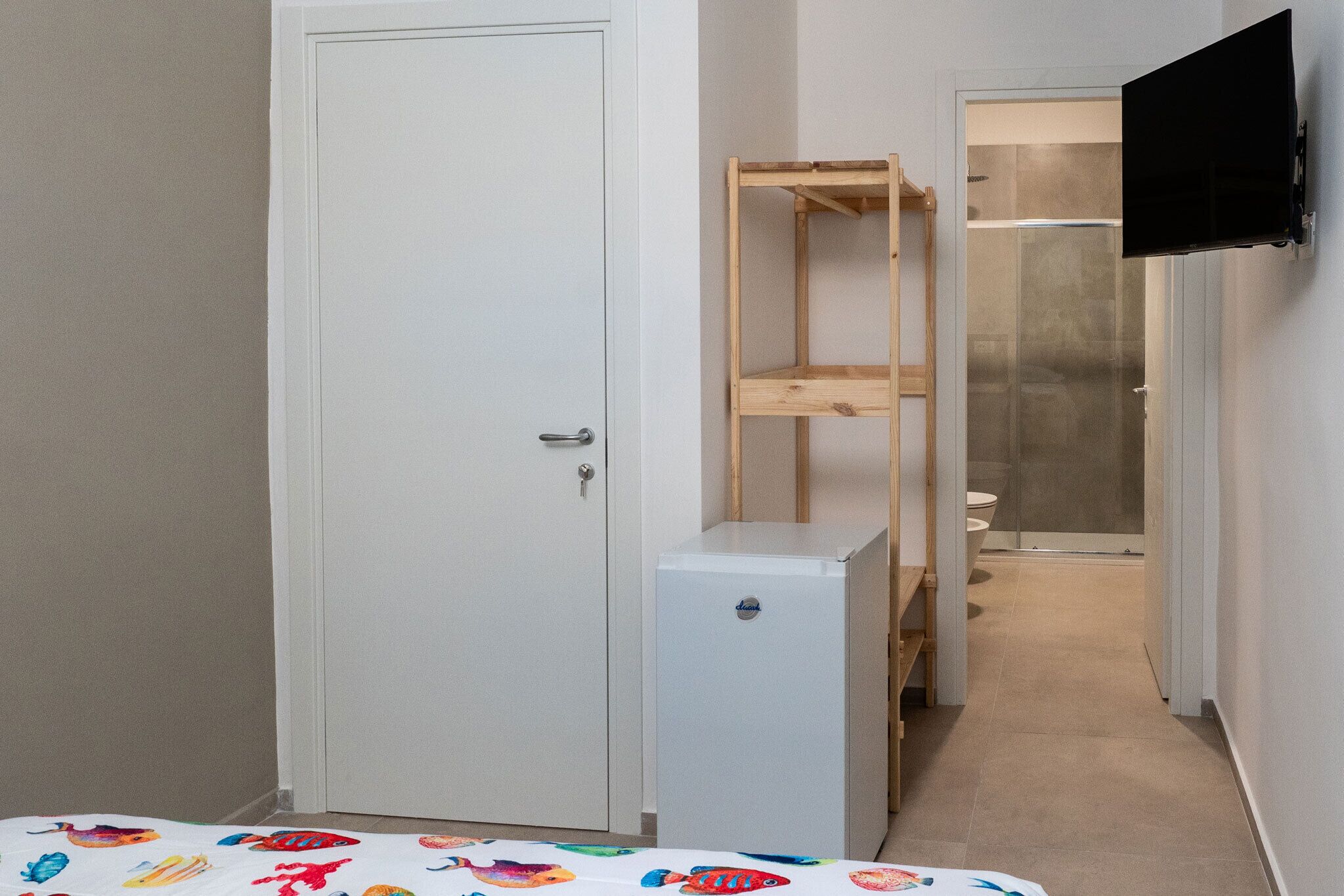 Un pasillo estrecho con una puerta blanca, un refrigerador pequeño de color blanco, una estantería de madera y un inodoro visible a través de un separador de vidrio.
