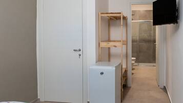 Un pasillo estrecho con una puerta blanca, un refrigerador pequeño de color blanco, una estantería de madera y un inodoro visible a través de un separador de vidrio.