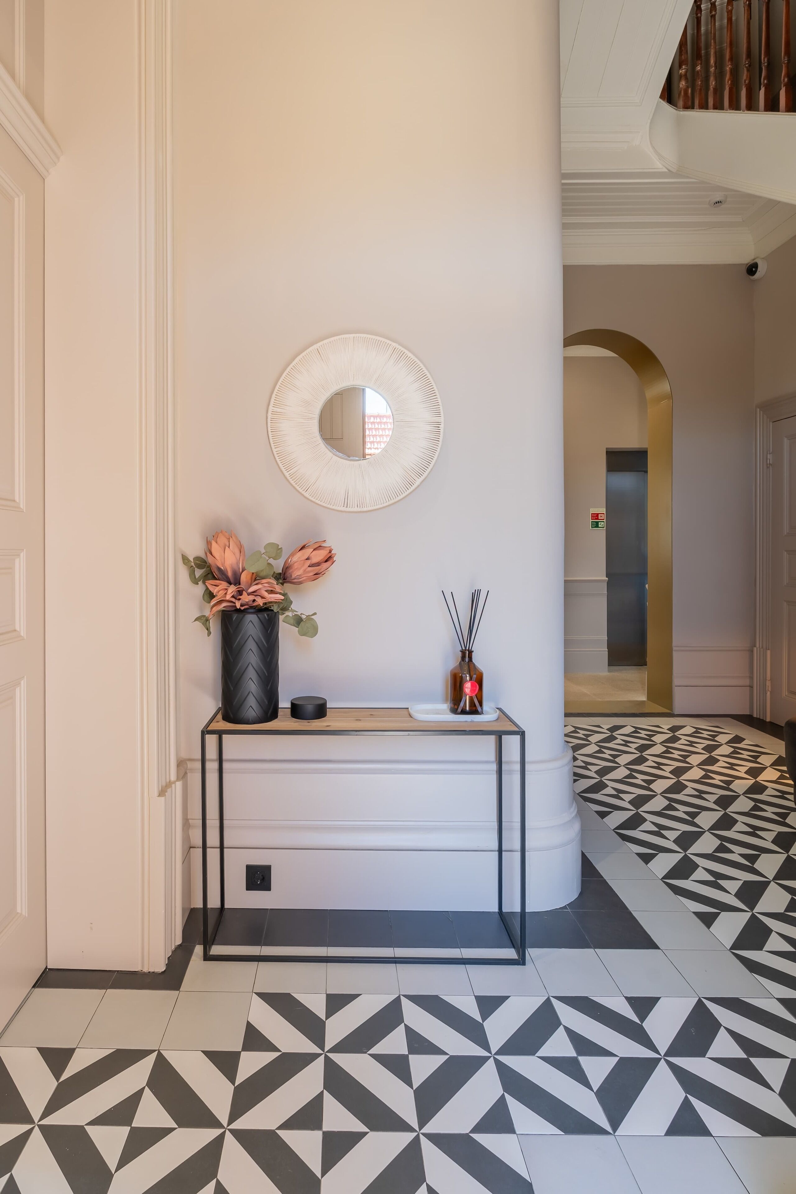 A hallway with a black and white patterned floor, a white console table with a vase of flowers, a mirror, and a door with a sign.