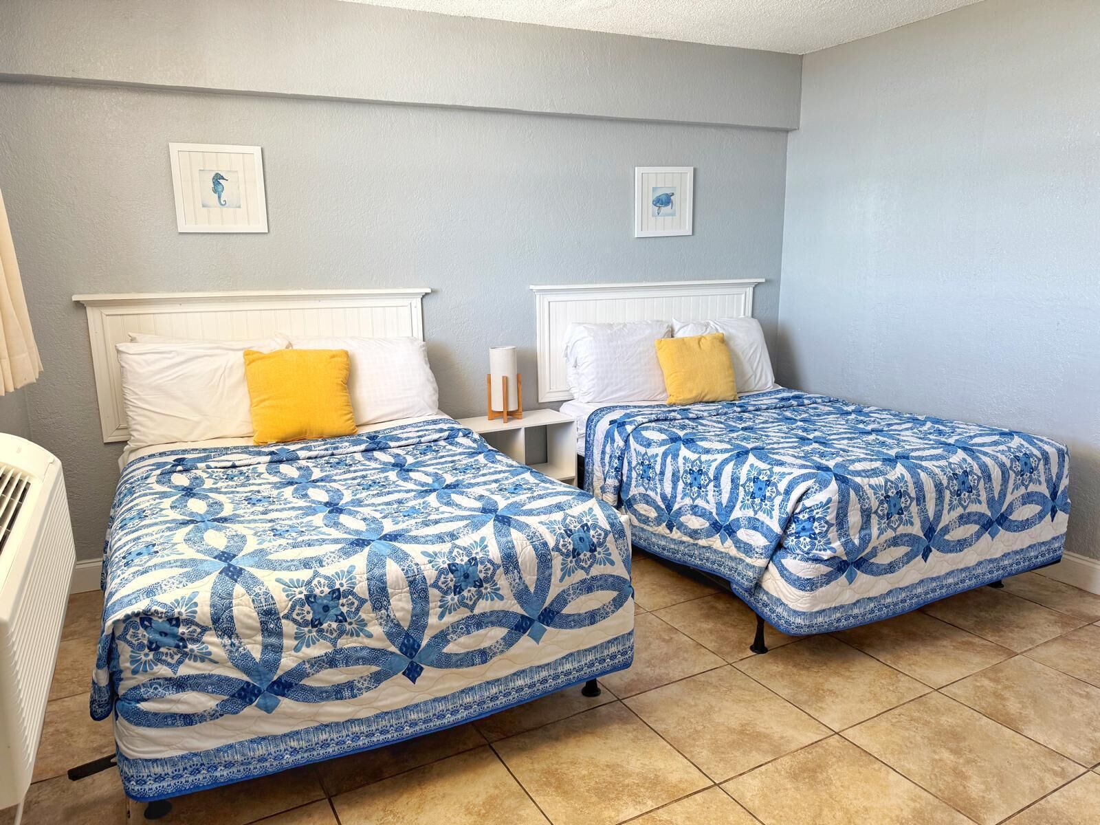 A room with two beds, each with a blue and white patterned bedspread, white headboards, and a small bedside table with a lamp.