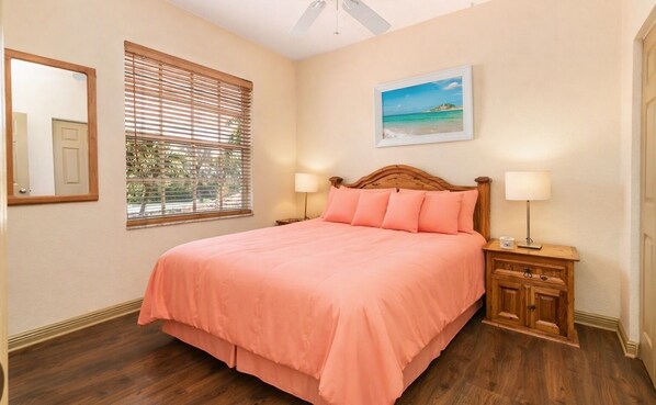 Um quarto com uma cama, duas mesas de cabeceira, um criado-mudo, uma janela com persianas, um quadro na parede e um ventilador de teto.