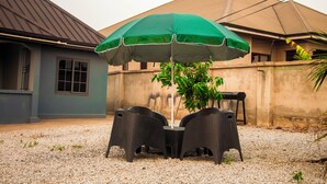 Un área de estar al aire libre con una mesa y sillas bajo un paraguas verde, rodeada de grava y un edificio con techo gris.