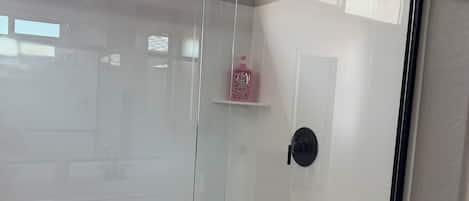 A glass-enclosed shower area with a black frame, a white door, and a pink bottle on a shelf.