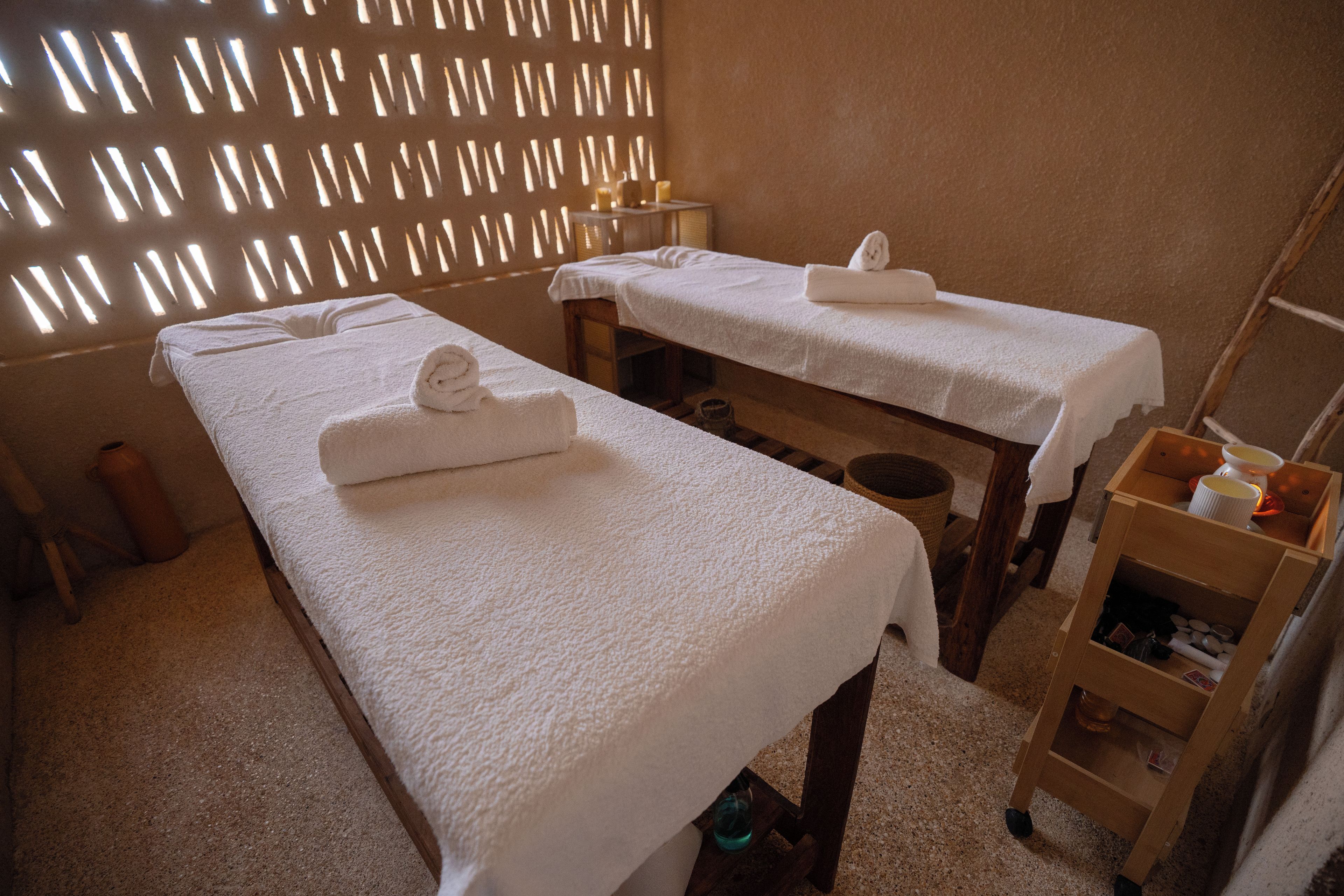 Une pièce comprenant deux tables de massage, chacune étant équipée d’une serviette et d’une bougie. On y trouve également une chaise en bois et une étagère avec des bouteilles.
