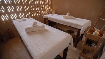 Een kamer met twee massagetafels, elk voorzien van een handdoek en een kaars. Er staat een houten stoel en een plank met flessen.