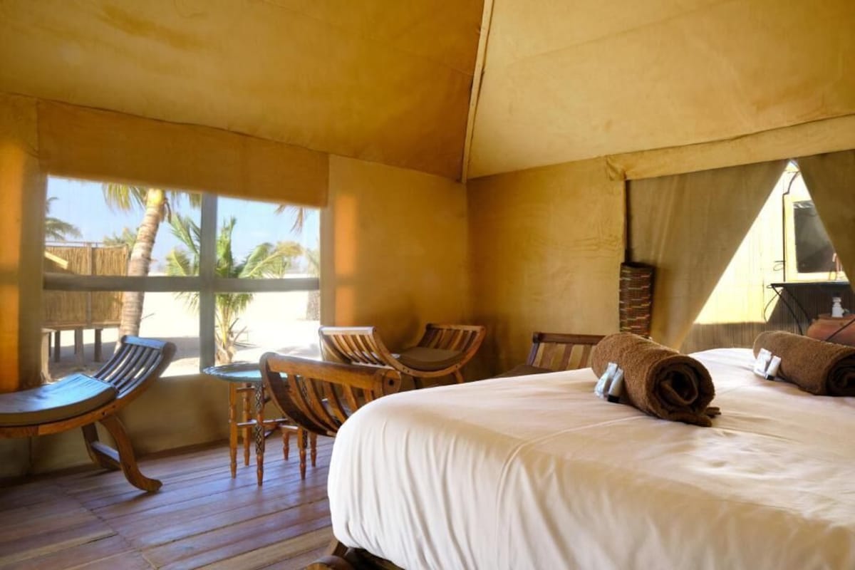 A room with a bed, wooden chairs, a small table, and a view of palm trees.