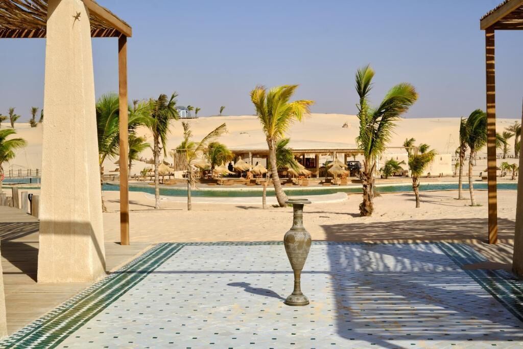 Ett semesterparadis vid stranden med palmer, en pool och en lusthusliknande byggnad.