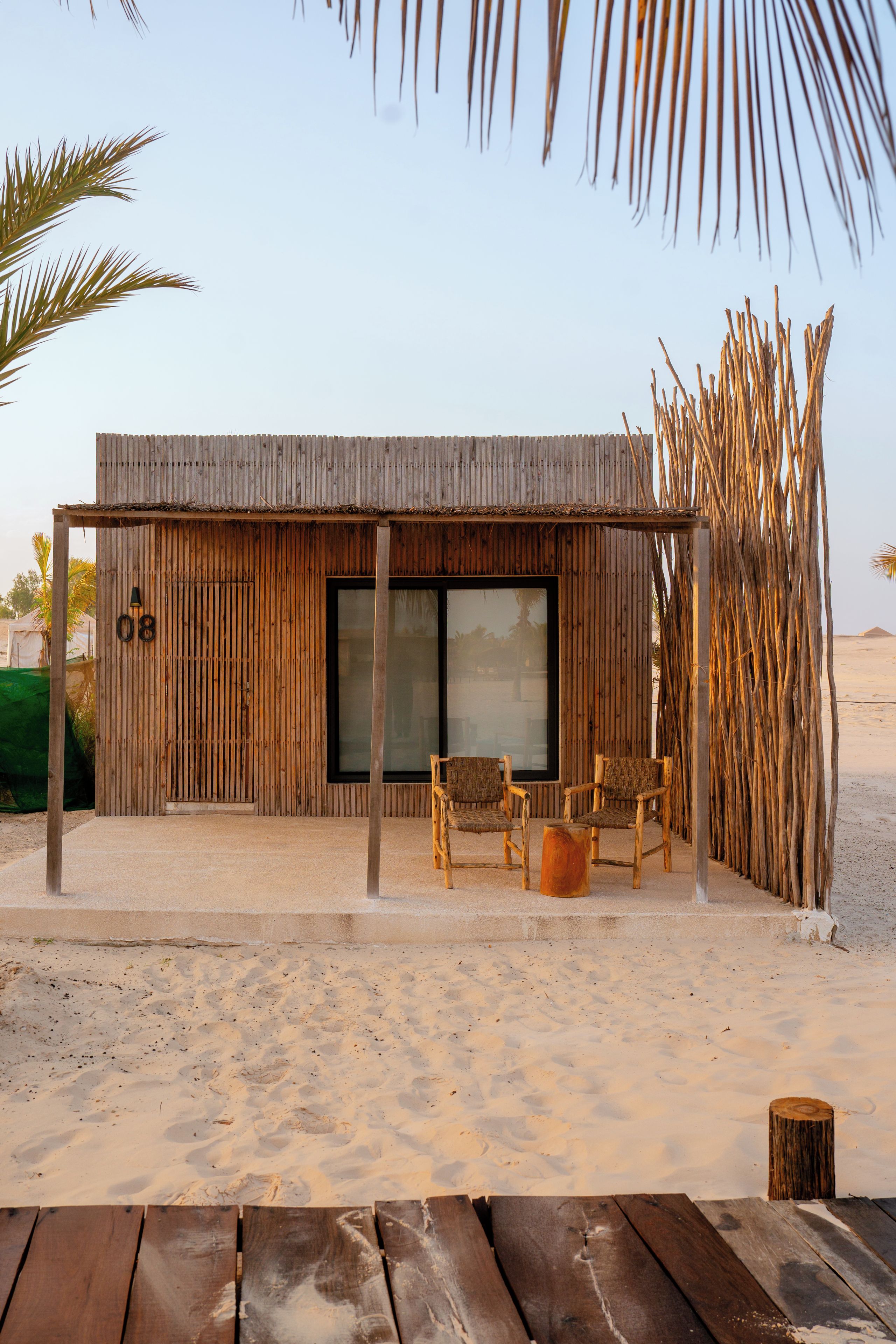 A wooden structure with a large window, a seating area with chairs, and a sandy ground.