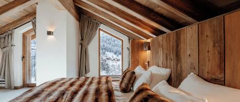 A bedroom with a wooden ceiling, a large window with a view of snow-covered trees, and a bed with white bedding.