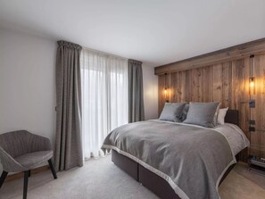 A bedroom with a bed, a chair, wooden panel walls, and a window with curtains. - DOMA Chalet l'Amont (Méribel)