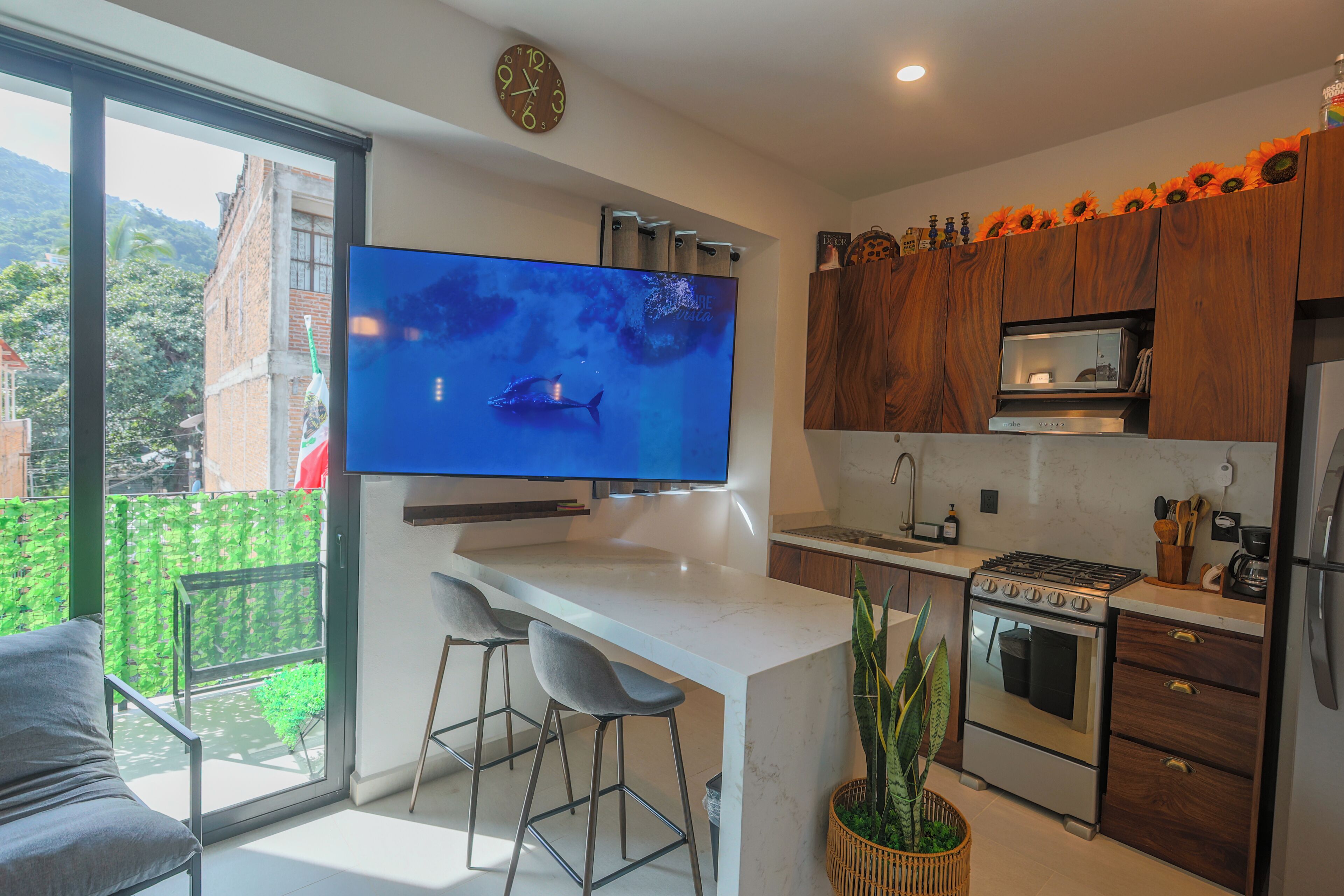 Una cocina moderna con una televisión de pantalla plana en la pared, una zona de comedor con taburetes de bar y un amplio ventanal con vistas al exterior.