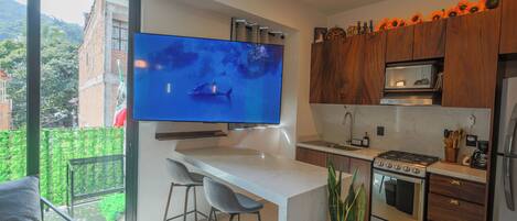 Una cocina moderna con una televisión de pantalla plana en la pared, una zona de comedor con taburetes de bar y un amplio ventanal con vistas al exterior.