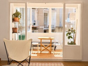 A room with a chair, a table, and a view through a glass door to an outdoor balcony with plants and a railing. - Grand T2 lumineux • Gare St-Charles & Vieux-Port (Marseille)