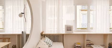 Une chambre à coucher au style minimaliste, avec un lit, une table de chevet, un miroir et une fenêtre avec des rideaux.