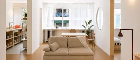 A modern living room with a sofa, a dining table, and a kitchen area visible through an open door.