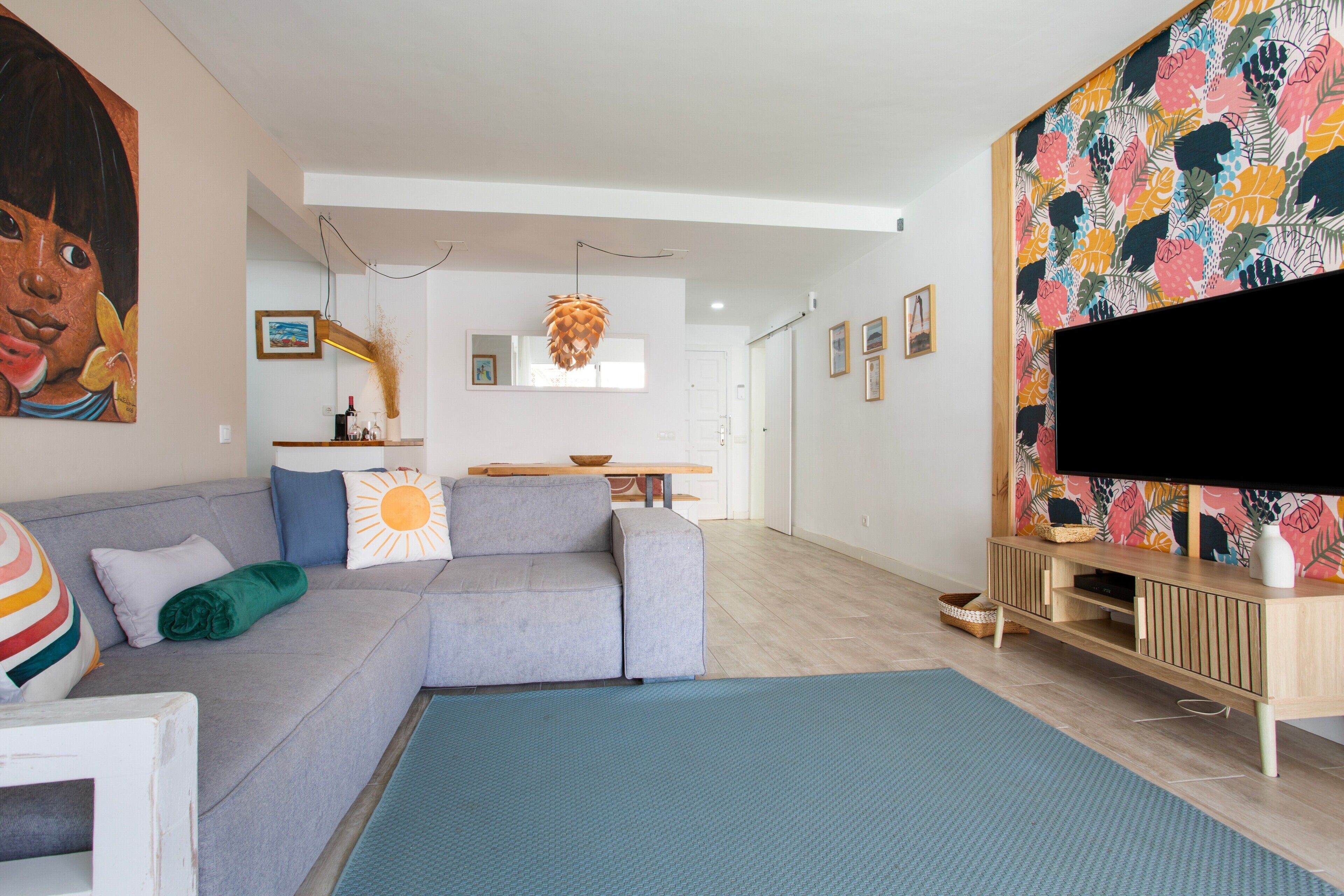 Een moderne woonkamer met een grijze bank, een flatscreen televisie en een kleurrijk muurdecor.