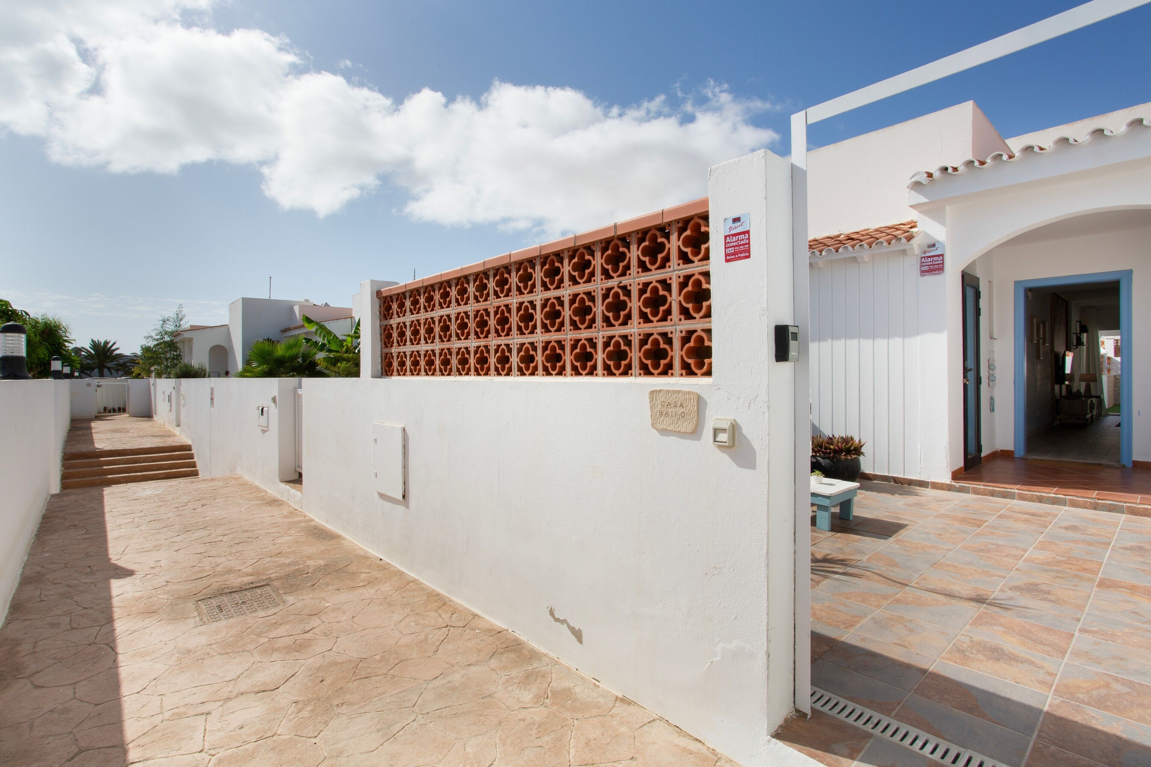 Un edificio blanco con un patio empedrado, una reja con diseño de celosía roja y un letrero que indica un sistema de alarma conectado.