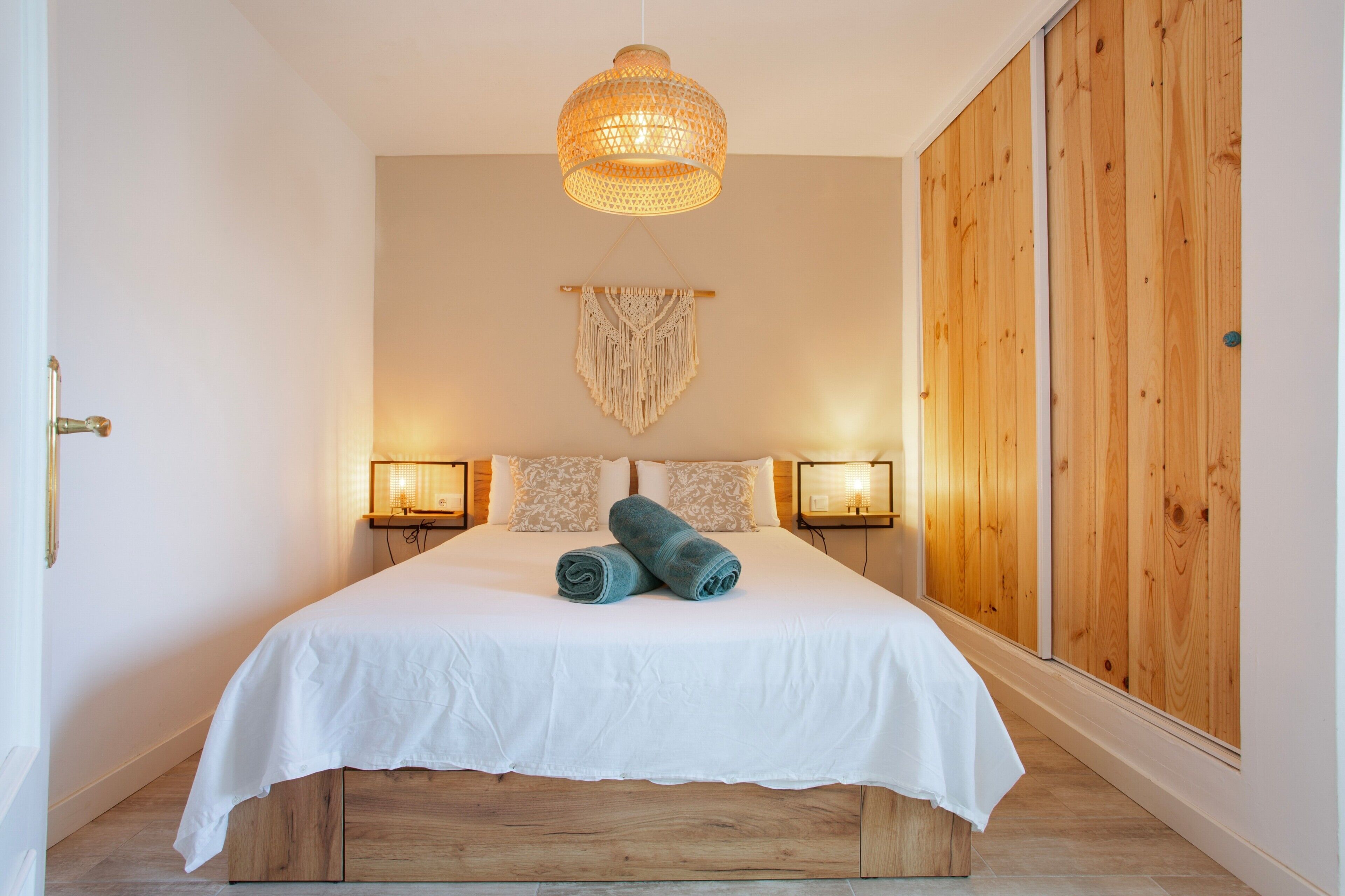 Een hotelkamer met een groot bed, een houten hoofdeinde, twee bedlampen, een macramé wanddecoratie en een houten kledingkast.