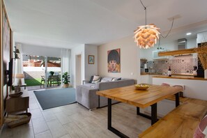 Uma sala de estar moderna com um sofá cinzento, uma mesa de jantar de madeira e uma área de cozinha com micro-ondas e painel decorativo.