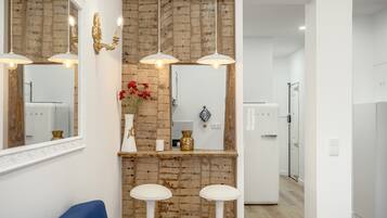 Une salle de bain moderne avec un mur d’accent en briques, un lavabo blanc et un miroir au-dessus. On y trouve deux tabourets blancs et une chaise bleue.