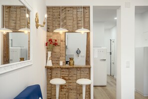 Un bagno moderno con una parete decorata in mattoni, un lavandino bianco e uno specchio sopra. Si vedono due sgabelli bianchi e una sedia blu.