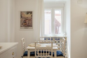 A room with a white table, a chair, a bed with a blue headboard, a window with a view of buildings, and a framed picture on the wall. - Bairro Alto Stylish II by Homing (Lisbon)