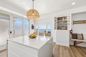 A modern kitchen with a large island, a hanging light fixture, and a view of the sea.