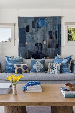 Een woonkamer met een groot blauw en zwart abstract wandtapijt, een bank met gekleurde kussens, een salontafel met boeken en een bloevas, en een raam met uitzicht op bomen.
