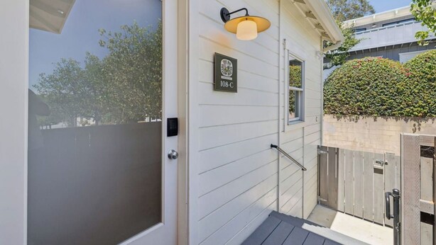 A white exterior with a door, a wall-mounted light fixture, and a hedge. - The Big Yellow House Lofts by the beach (Summerland)