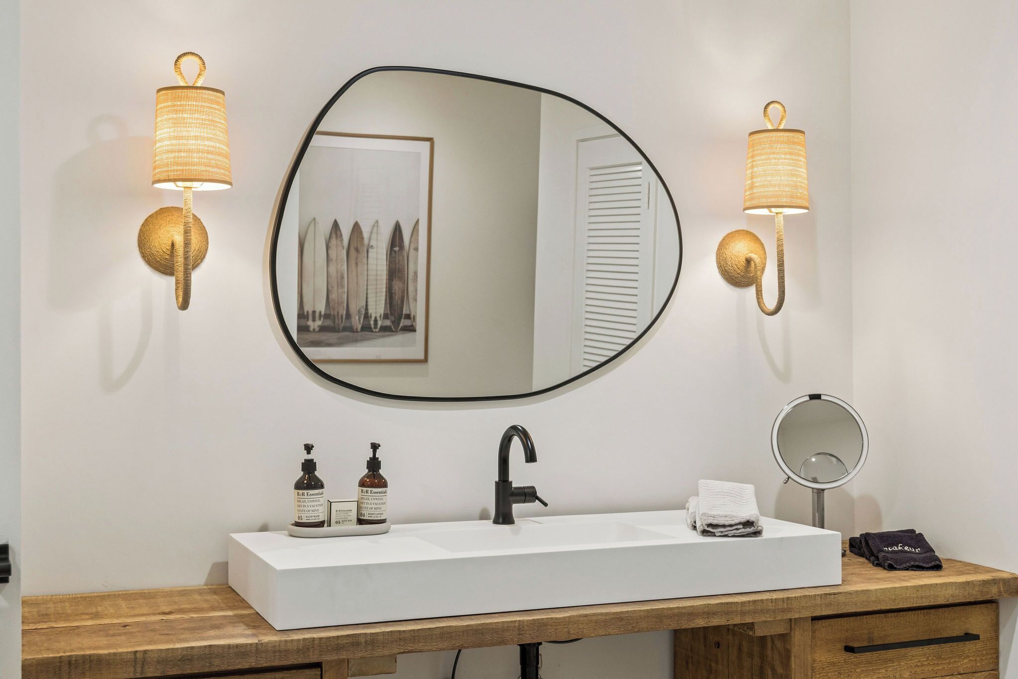 A bathroom with a white sink, black faucet, two bottles of R&R Essential hand wash, a round mirror, and two wall-mounted lamps.