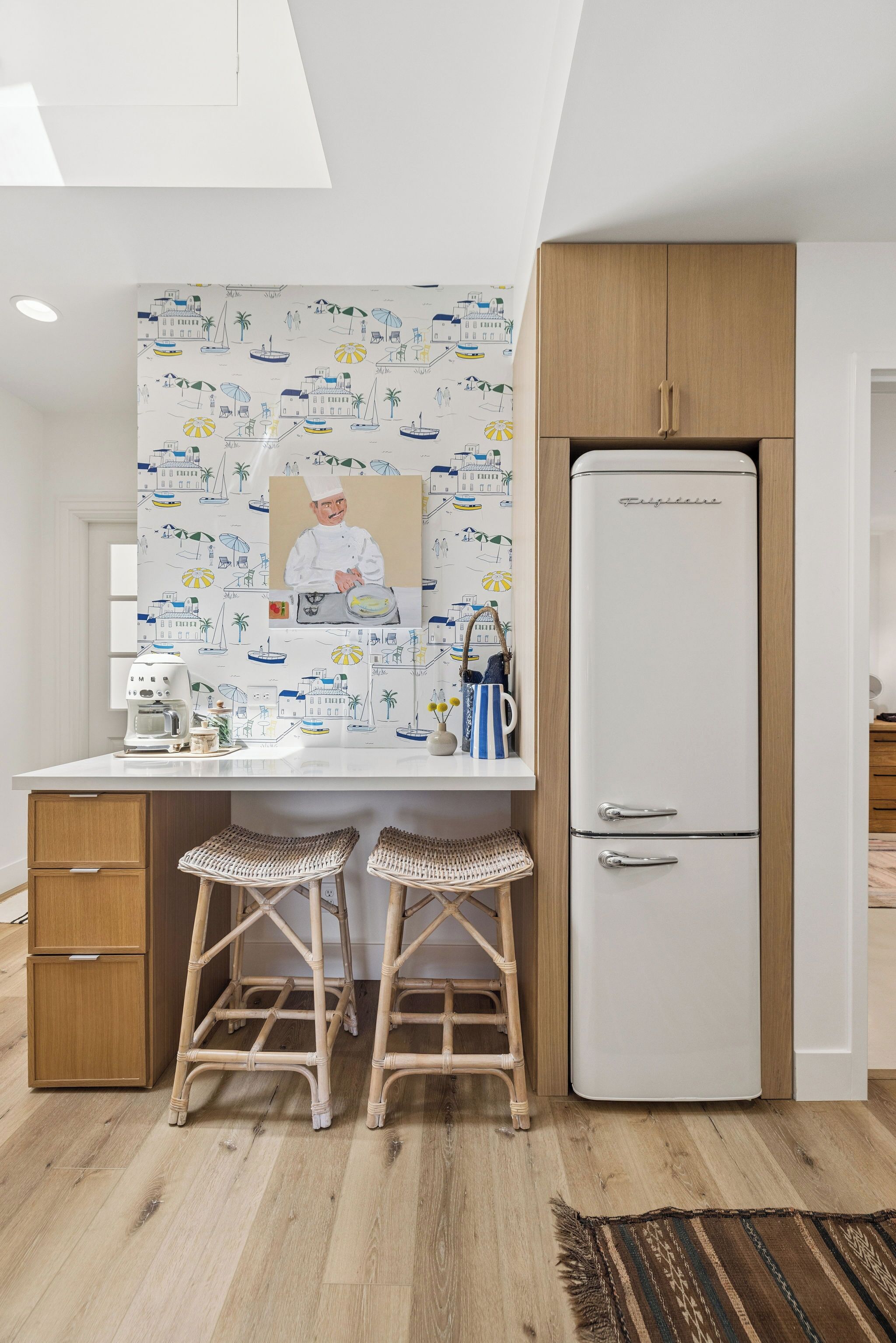 A kitchen with a white refrigerator, wooden cabinets, a white countertop, two wicker stools, and a wall with a beach-themed mural.