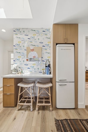 Een keuken met een witte koelkast, houten kastjes, een wit aanrecht, twee rieten krukken en een muur met een strandthema-muurschildering.