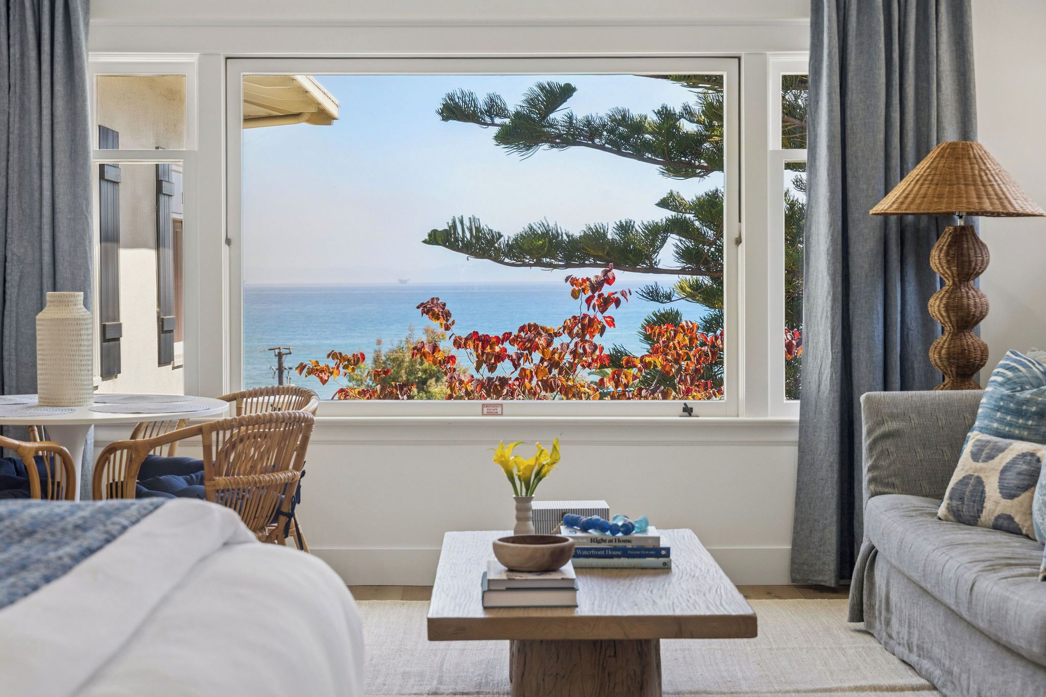 A room with a view of the sea, a window with a view of trees and flowers, a table with books and a vase, and a lamp.