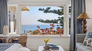 A room with a view of the sea, a window with a view of trees and flowers, a table with books and a vase, and a lamp. - The Big Yellow House Lofts by the beach (Summerland)