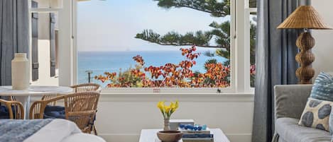 Una habitación con vistas al mar, una ventana que da a árboles y flores, una mesa con libros y un jarrón, y una lámpara.