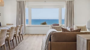 A room with a large window, a dining area with wicker chairs, a sofa, and a view of the sea. - The Big Yellow House Lofts by the beach (Summerland)