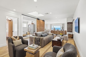 Uma sala de estar moderna com um sofá, poltronas e uma mesa de centro. É visível, ao fundo, uma área de jantar com mesa e cadeiras.