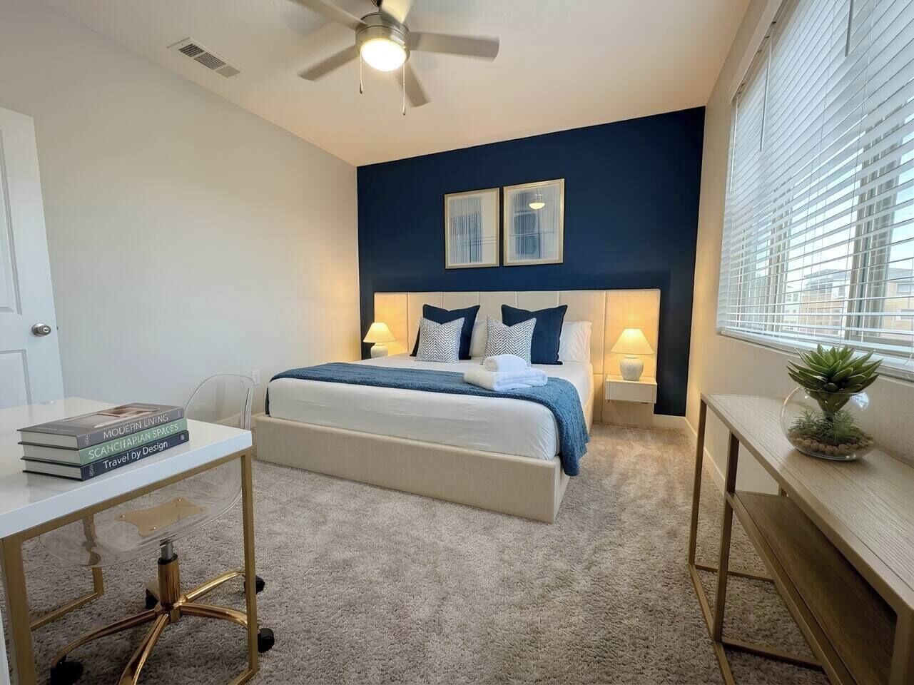 Une chambre à coucher comprenant un lit, des tables de chevet, un bureau, un ventilateur de plafond et une fenêtre avec des stores.