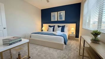 Une chambre à coucher comprenant un lit, des tables de chevet, un bureau, un ventilateur de plafond et une fenêtre avec des stores.