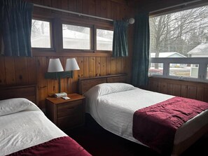 Una habitación con dos camas, paredes de madera, una ventana con cortinas y una mesita de noche con lámparas.