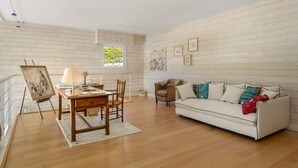 Uma sala de estar ampla com piso de madeira, um sofá branco, uma escrivaninha de madeira com uma luminária e um quadro na parede.