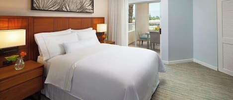 A hotel room with a large bed, a nightstand with a lamp, a wall-mounted artwork, a sliding door to a balcony, and a view of the outdoors.