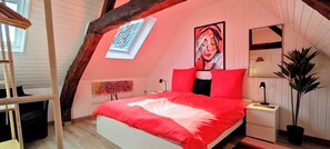 A bedroom with a sloped ceiling, a bed with red bedding, a large mirror, and a potted plant.